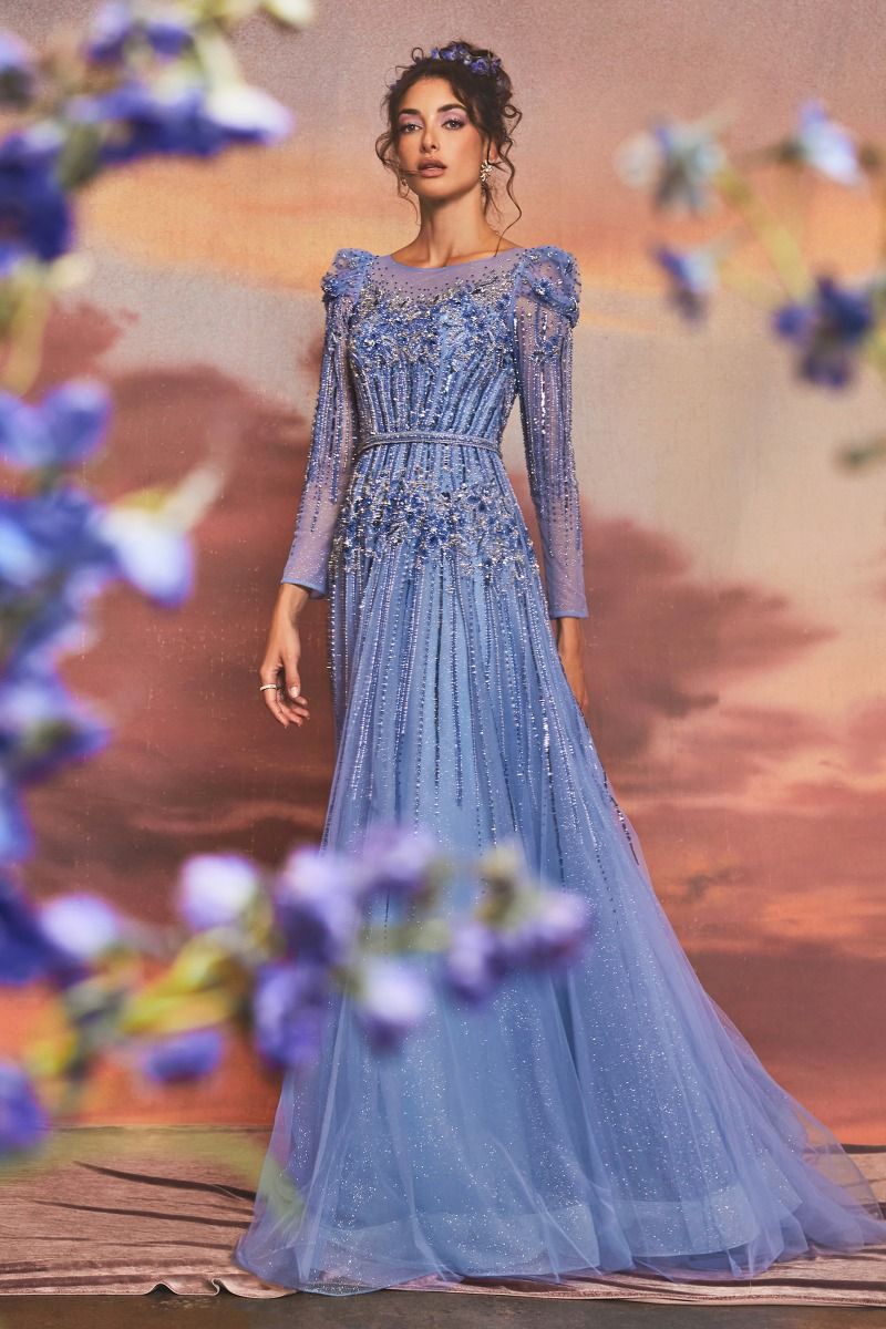 A woman stands before a sunset backdrop wearing the Luxurious Formal Gown A1167 by Andrea and Leo, featuring long sleeves and sheer, embellished floral details; purple flowers bloom in the foreground.
